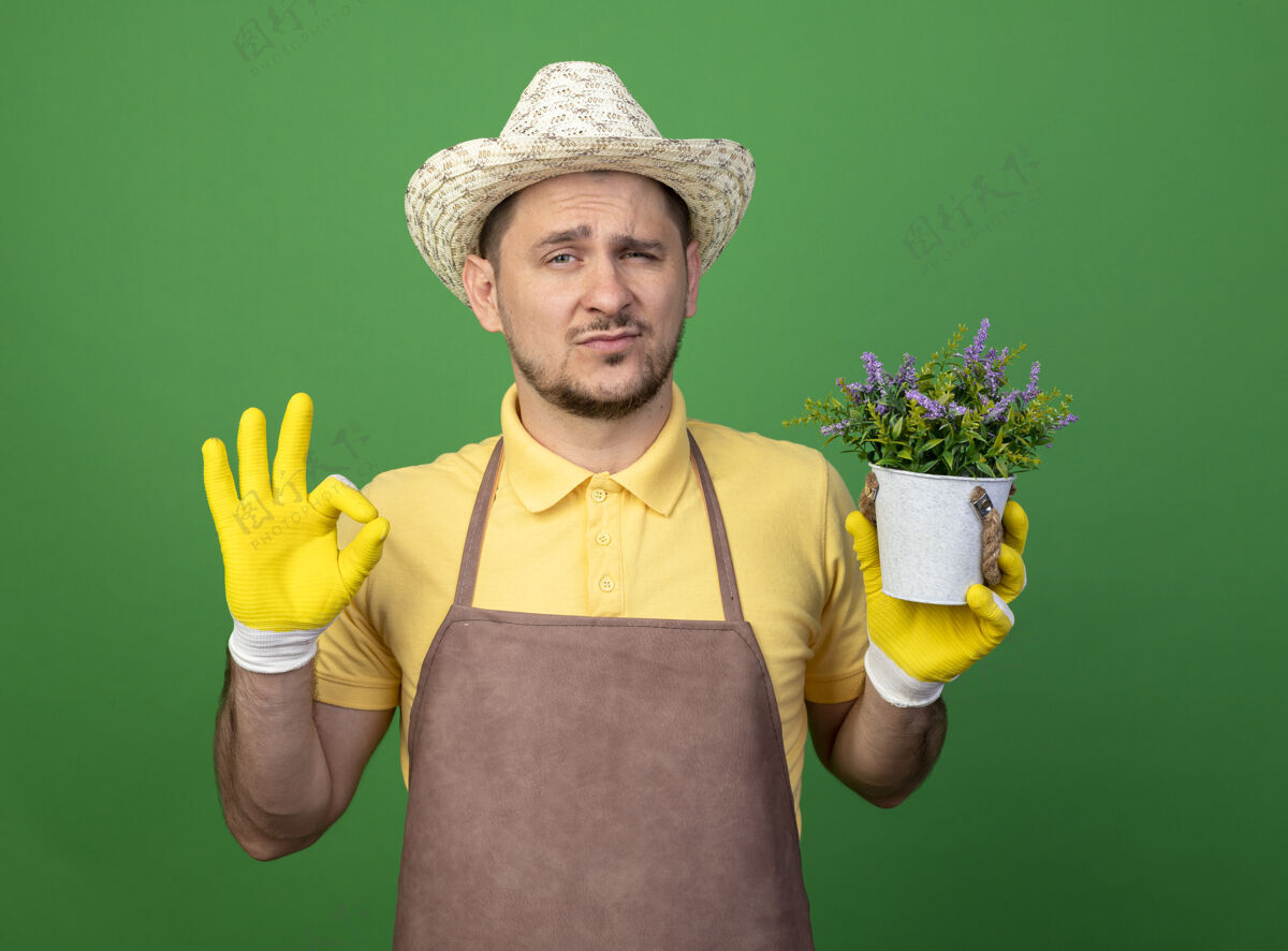 看年轻的园丁穿着连体衣 戴着帽子 戴着工作手套 手里拿着盆栽植物 自信地看着前面 绿色的墙上挂着“ok”的牌子信心人人