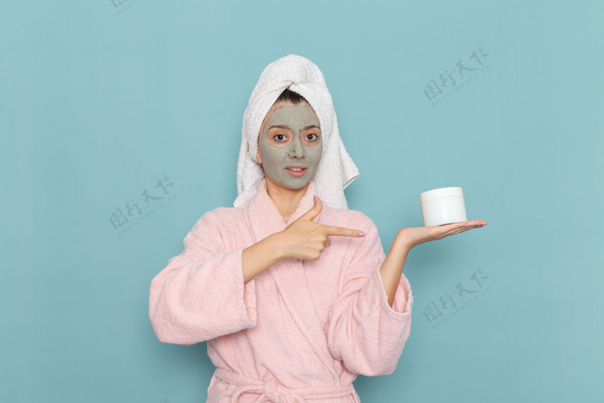 面部正面图：年轻女性 穿着粉色浴衣 脸上戴着口罩 拿着淡蓝色的面霜 淋浴 清洁 美容 自我护理年轻人成人面具