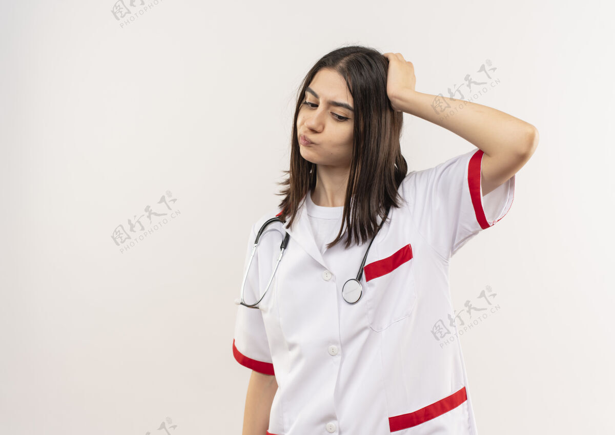 旁白年轻的女医生穿着白大褂 脖子上戴着听诊器 站在白墙边茫然地看着一边姿势拼图人
