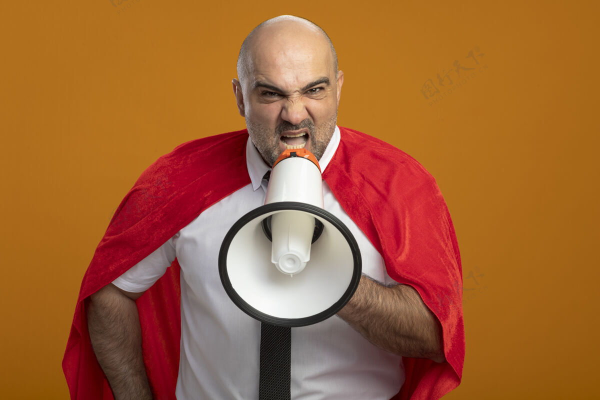 侵略性愤怒的超级英雄商人穿着红色斗篷站在橙色的墙上对着扩音器大喊大叫 表情咄咄逼人超级英雄商人表情