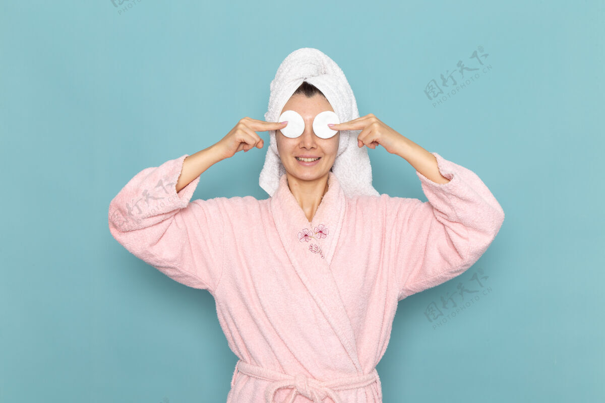 年轻正面图身着粉色浴袍的年轻女性在蓝色墙上用小棉布遮住眼睛淋浴清洁美容自护霜棉花自我照顾前面