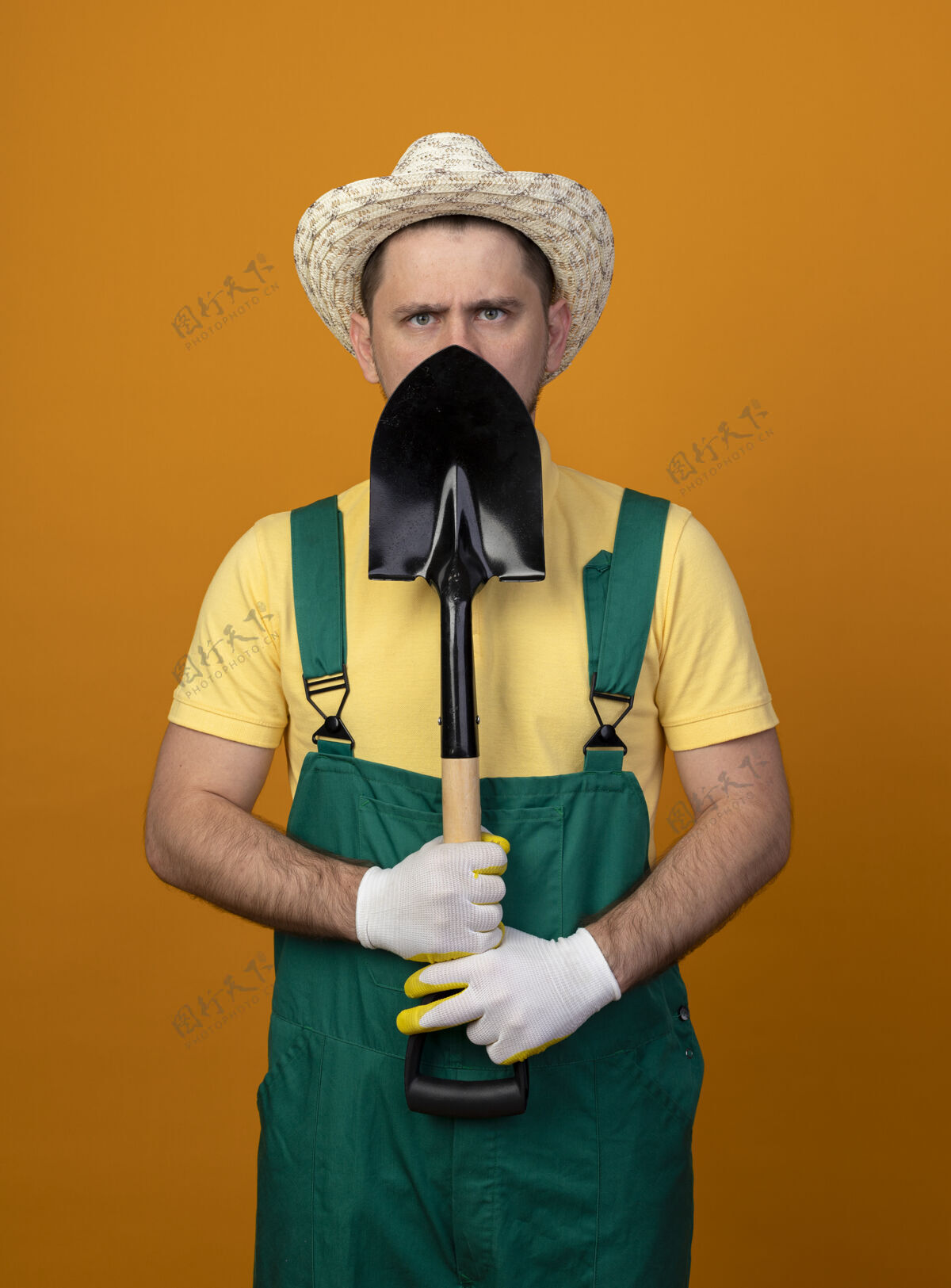 站着年轻的园丁穿着连体衣 拿着铲子 严肃的脸站在橙色的墙上 看着前面橙色工作帽子