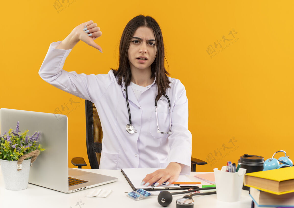 年轻人身穿白大褂 手持听诊器的年轻女医生看起来很不高兴 大拇指朝下坐在桌子旁 笔记本电脑和文件放在橙色的墙上笔记本电脑羽绒服人