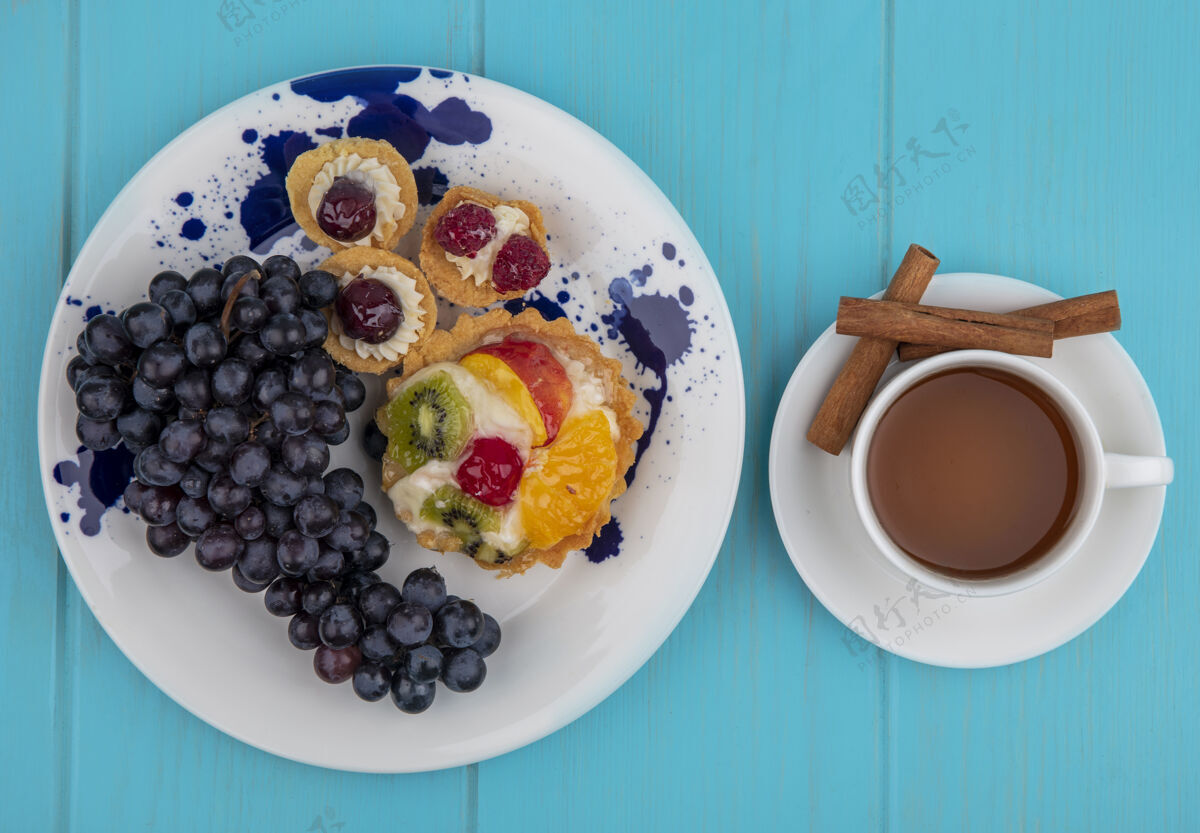 景观水果蛋糕和葡萄在盘子里的俯视图 以及一杯茶和肉桂茶碟在蓝色背景上的俯视图茶甜点肉桂