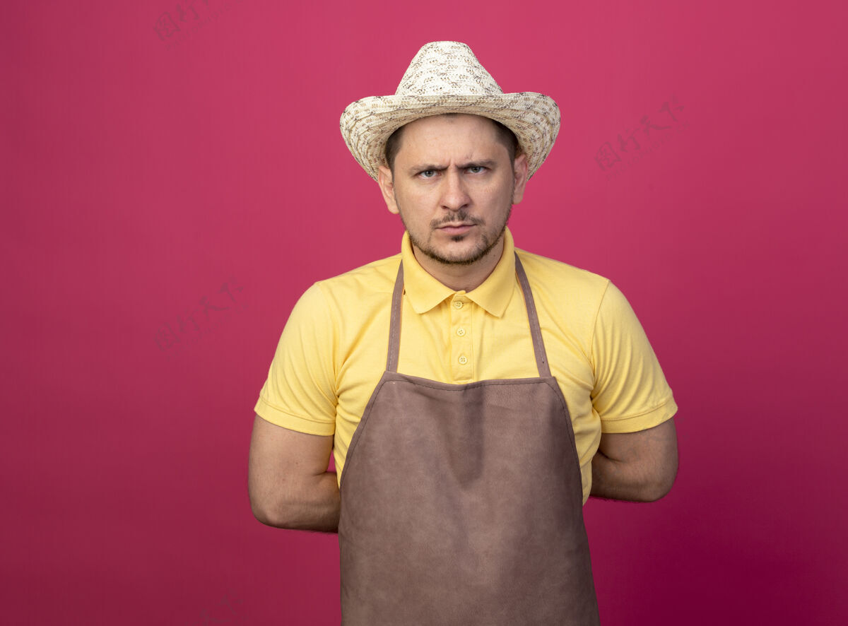 男人年轻的园丁穿着连体衣 戴着帽子 戴着工作手套 站在粉红色的墙上严肃地看着前面 脸上露出不高兴的表情严重脸花园