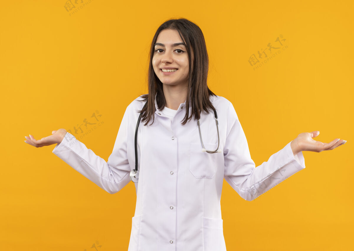 站年轻的女医生穿着白大褂 脖子上戴着听诊器 困惑地看着前方 微笑着张开双臂站在橙色的墙上医疗保健微笑外套