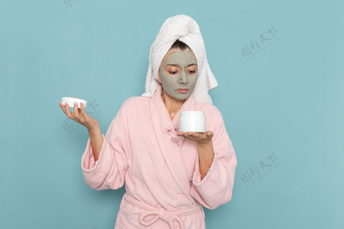 前面正面图身着粉色浴袍的年轻女性拿着面霜罐在蓝色墙上淋浴清洗美容自护面霜人浴袍脸