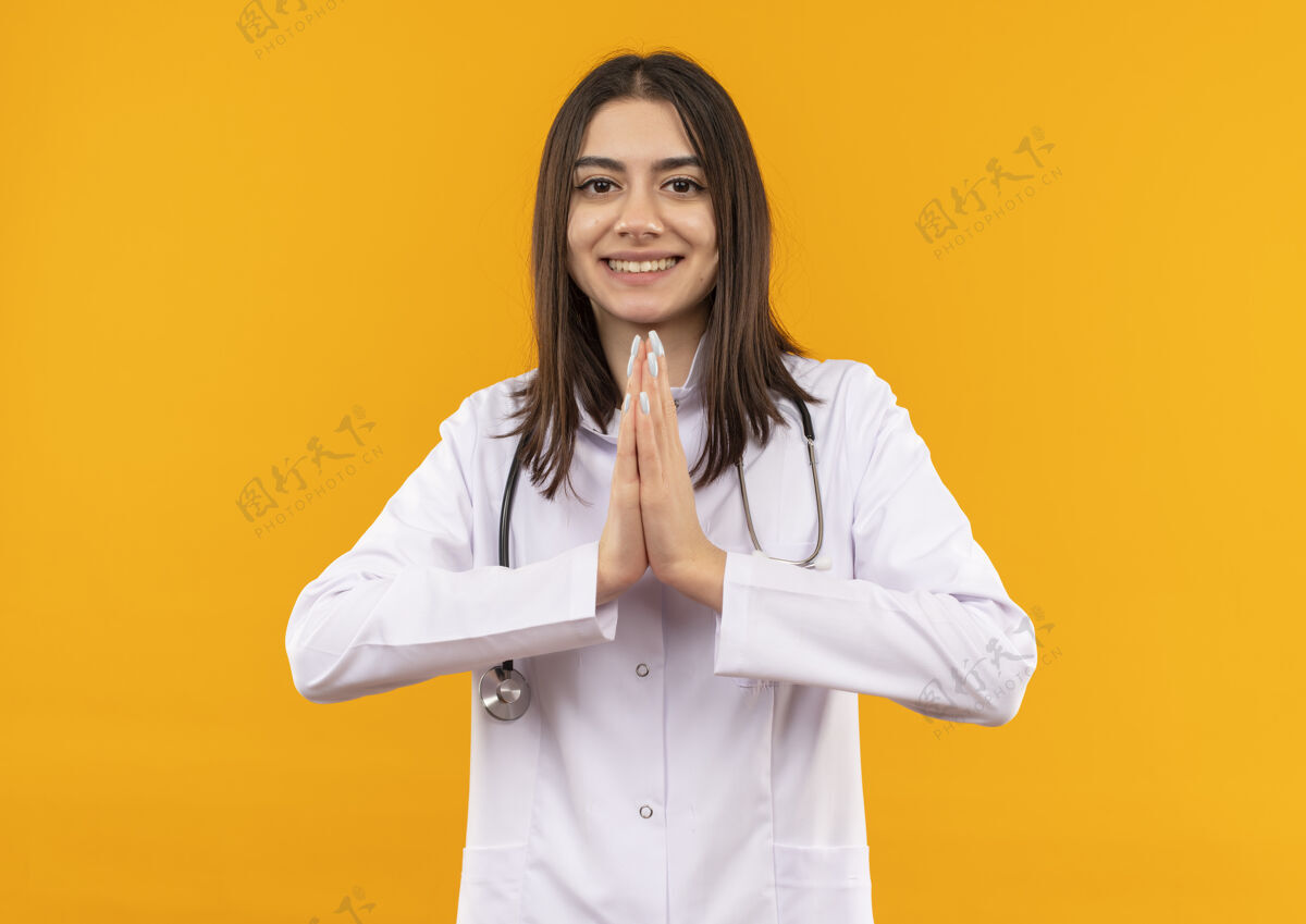 医生身穿白大褂 脖子上戴着听诊器的年轻女医生望着前方 双手合十 站在橘红色的墙上 露出感激的微笑纳马斯特看着在身边