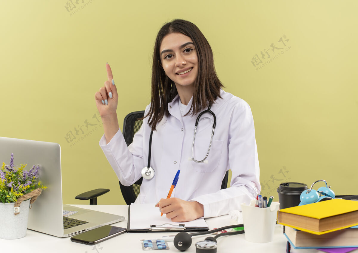 新的年轻的女医生穿着白大褂 脖子上戴着听诊器 食指有了新的想法坐在桌边 笔记本电脑放在墙上女性桌子外套