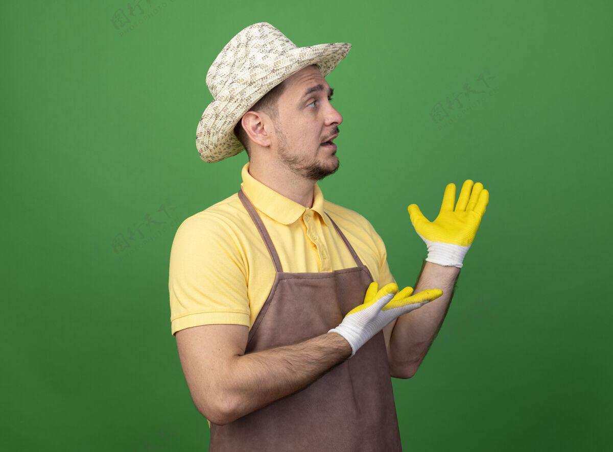 手套年轻的园丁穿着连体衣 戴着帽子 戴着工作手套 站在绿色的墙壁上 双手叉开 困惑地看着一边看困惑帽子