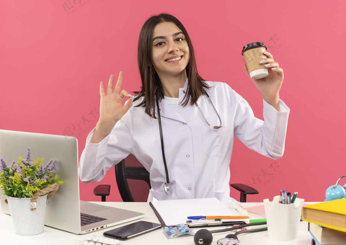 杯子年轻的女医生穿着白大褂 脖子上戴着听诊器 手里拿着一个咖啡杯 上面写着“ok”字样 微笑着坐在桌旁 笔记本电脑放在粉色的墙上市民外套办公室