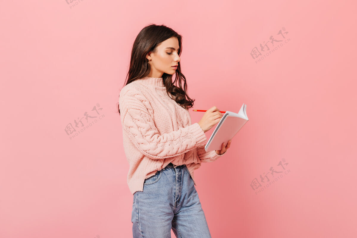 女孩卷发女孩专心地在日记里做笔记穿着毛衣的女人在粉色背景上摆姿势头发蓝眼睛笔记本
