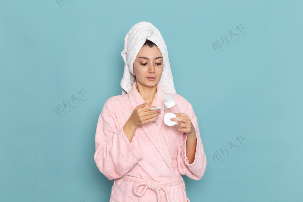 视图正面图年轻女性穿着粉色浴袍淋浴后在浅蓝色墙面上清洗美容洁面水自护霜淋浴脸年轻帽子