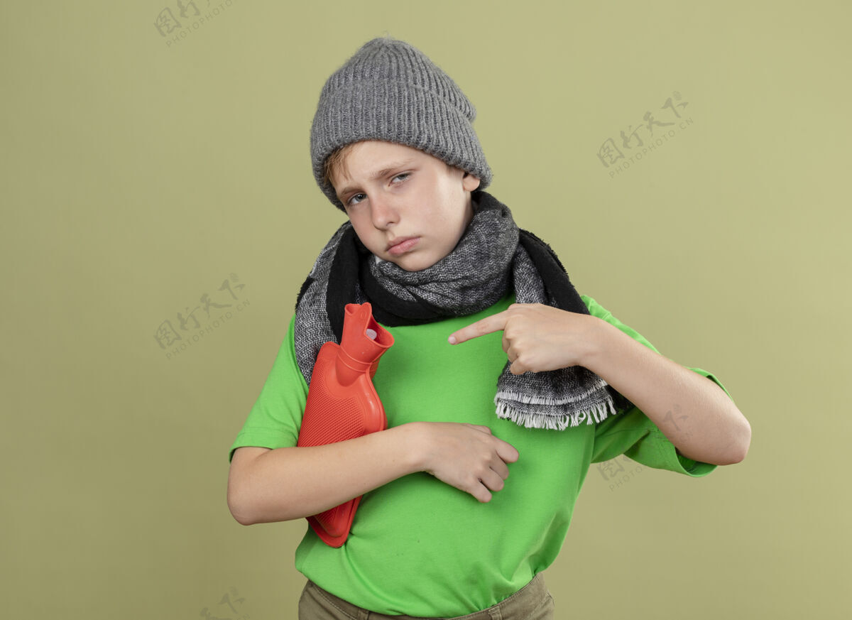 围巾生病的小男孩穿着绿色t恤 戴着暖和的围巾和帽子 感觉不舒服 拿着水瓶取暖 手指指着它站在明亮的墙上疾病保持指向