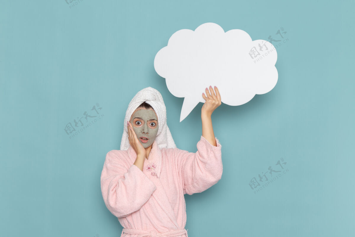 肖像正面图身着粉色浴袍的年轻女性手持蓝色墙壁上的白色大标牌清洁美容自护霜淋浴奶油浴袍查看