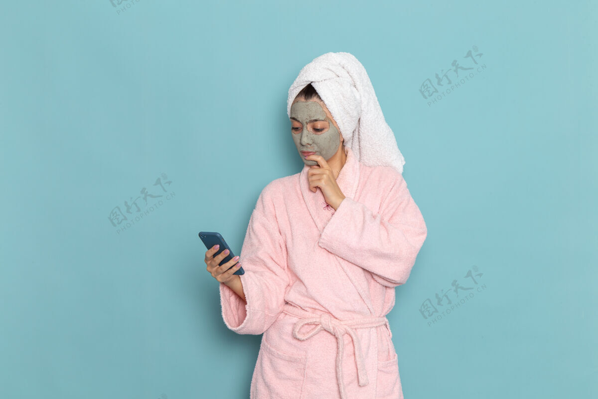 视图正面图年轻女性穿着粉色浴袍淋浴后用手机在浅蓝色墙面上美容水自理淋浴干净实验室外套淋浴浴袍