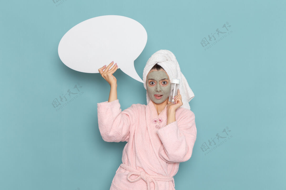 微笑正面图年轻女性穿着粉色浴袍淋浴后手持白色标牌 喷在蓝色墙面上的美容水霜自护淋浴浴袍肖像自我照顾