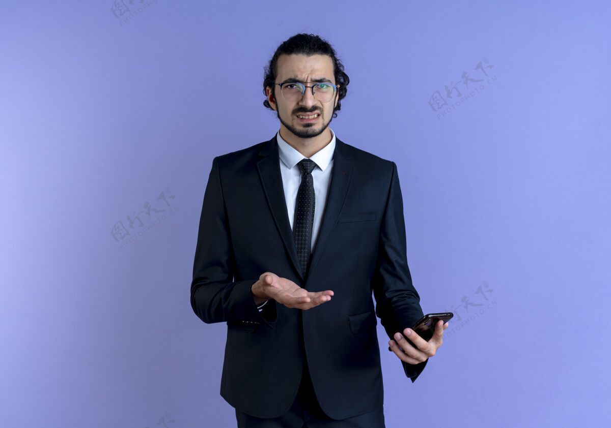 困惑一个穿着黑色西装戴着眼镜的商人拿着智能手机站在蓝色的墙上 困惑而不安地看着前方手机人看着