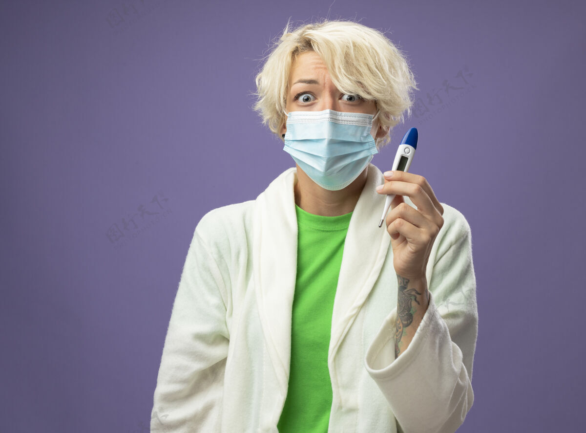 不健康生病的不健康的女人 戴着护面面具的短发 手里拿着体温计 心烦意乱地站在紫色的墙上女人体温计抱着