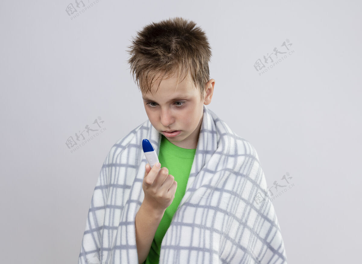 小生病的小男孩穿着绿色t恤 裹着毛毯 看着温度计 不安地站在白色的墙上看病担心