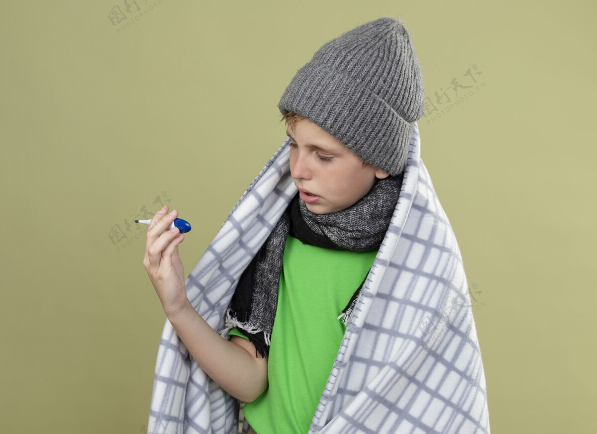 毯子生病的小男孩穿着绿色t恤 戴着暖和的围巾和帽子 裹着毯子 手里拿着温度计 站在光墙上焦急地看着它站包穿