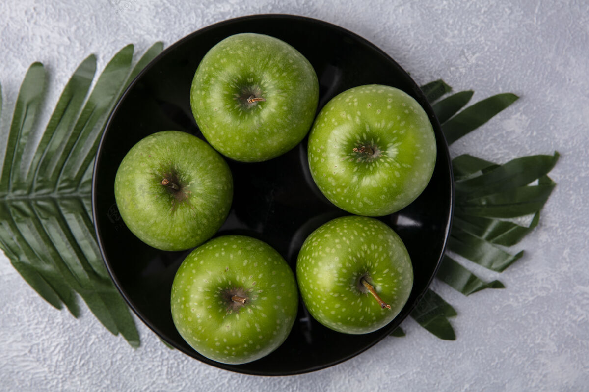 食物俯视图绿色的苹果在一个黑色的盘子上 树枝上的叶子在白色的背景上新鲜顶部绿色