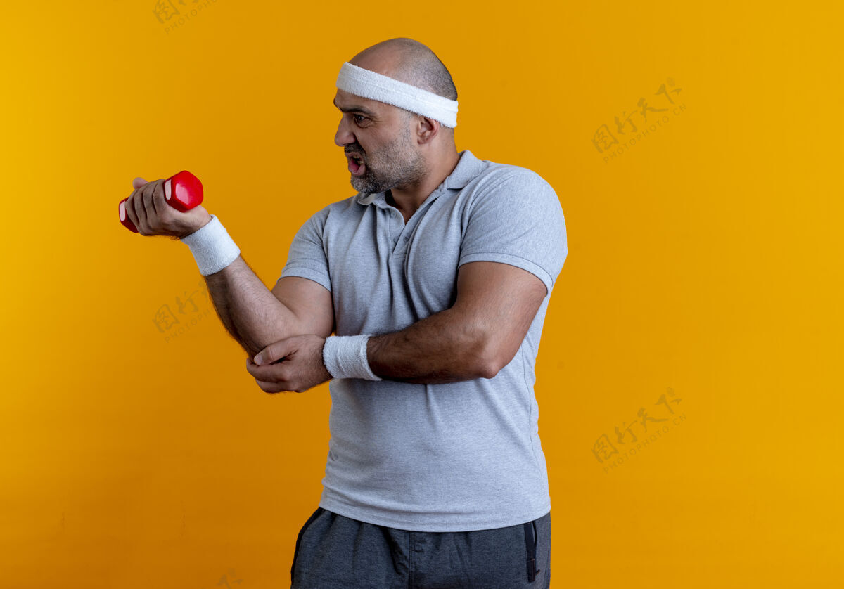 头带成熟的运动型男人戴着头巾 站在橘色的墙上 哑铃看起来很疲惫 精疲力尽姿势人哑铃
