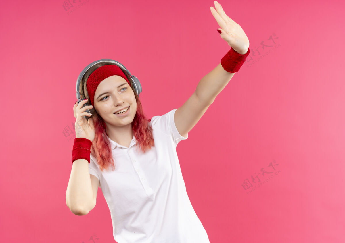 头带戴着耳机 戴着头带的年轻运动型女人一边看着一边 一边开心地挥舞着积极的手 站在粉色的墙上看站积极
