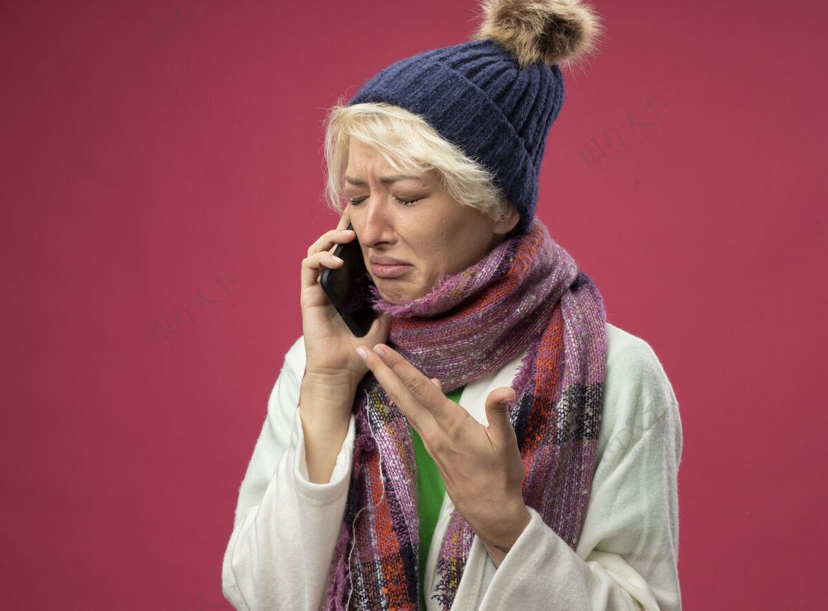 头发不舒服的生病的不健康的女人 短头发 戴着暖和的围巾和帽子 站在粉红色的墙上一边打电话一边哭不适短裤女人