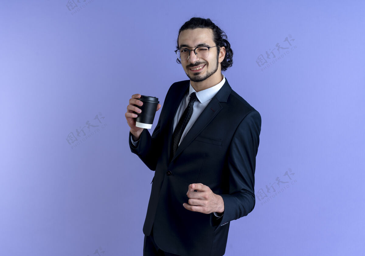 人一个穿着黑西装戴着眼镜的商人站在蓝色的墙上 手里拿着咖啡杯 握紧拳头 开心而积极地微笑着 眨着眼睛拳头快乐握紧