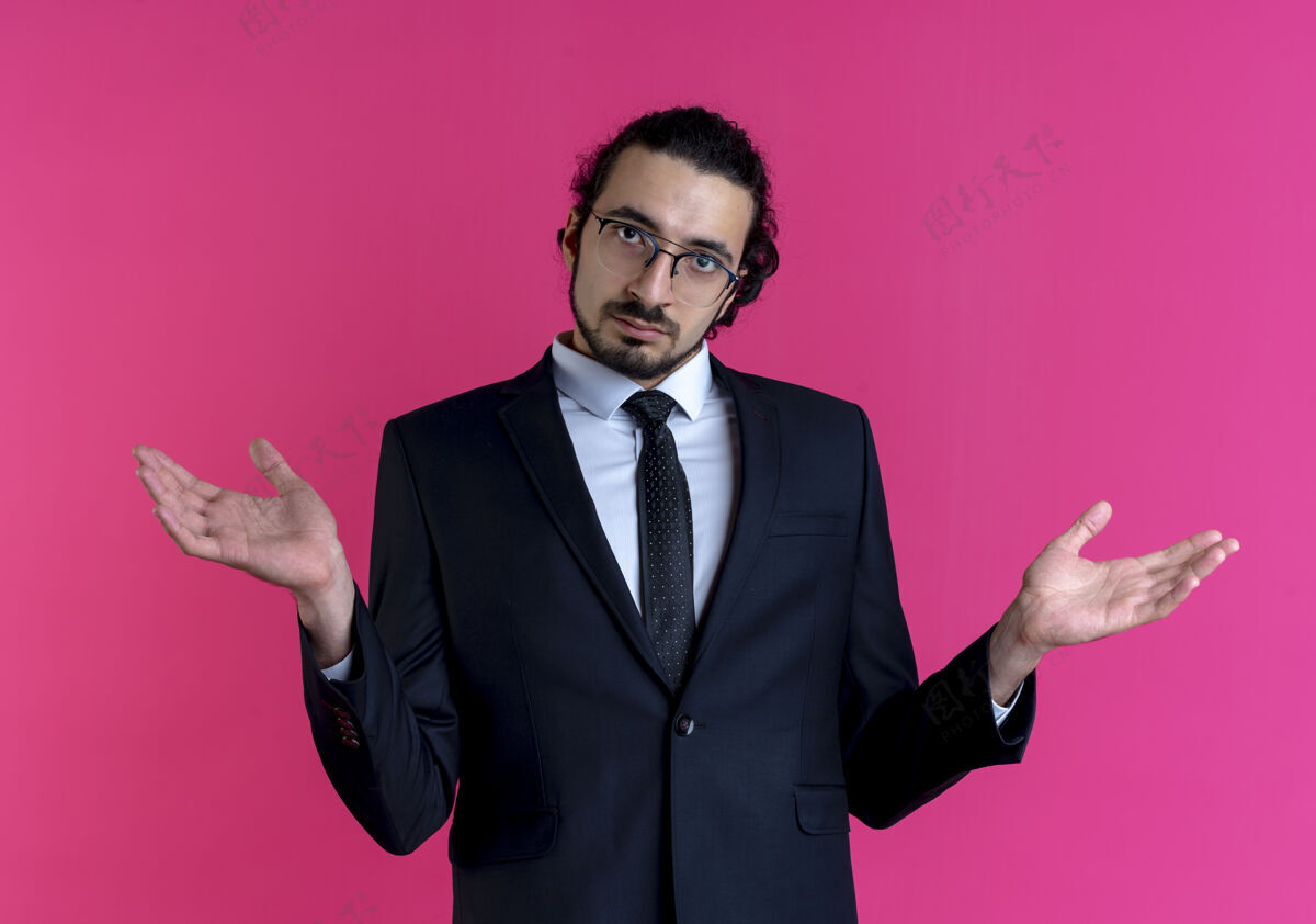 提高身穿黑色西装 戴着眼镜的商人站在粉红色的墙上 抬起双手 表情困惑地看着前方公民姿势手
