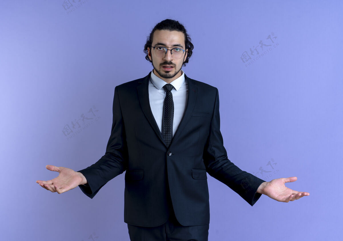 家伙一个穿着黑西装戴着眼镜的商人站在蓝色的墙壁上 看上去困惑而不确定地向两边伸出双臂领带看困惑