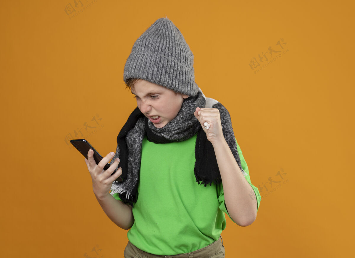 围巾生病的小男孩穿着绿色t恤 戴着暖和的围巾和帽子 感觉不舒服 拿着智能手机 站在橙色的墙上生气地看着它男孩可移动橙色