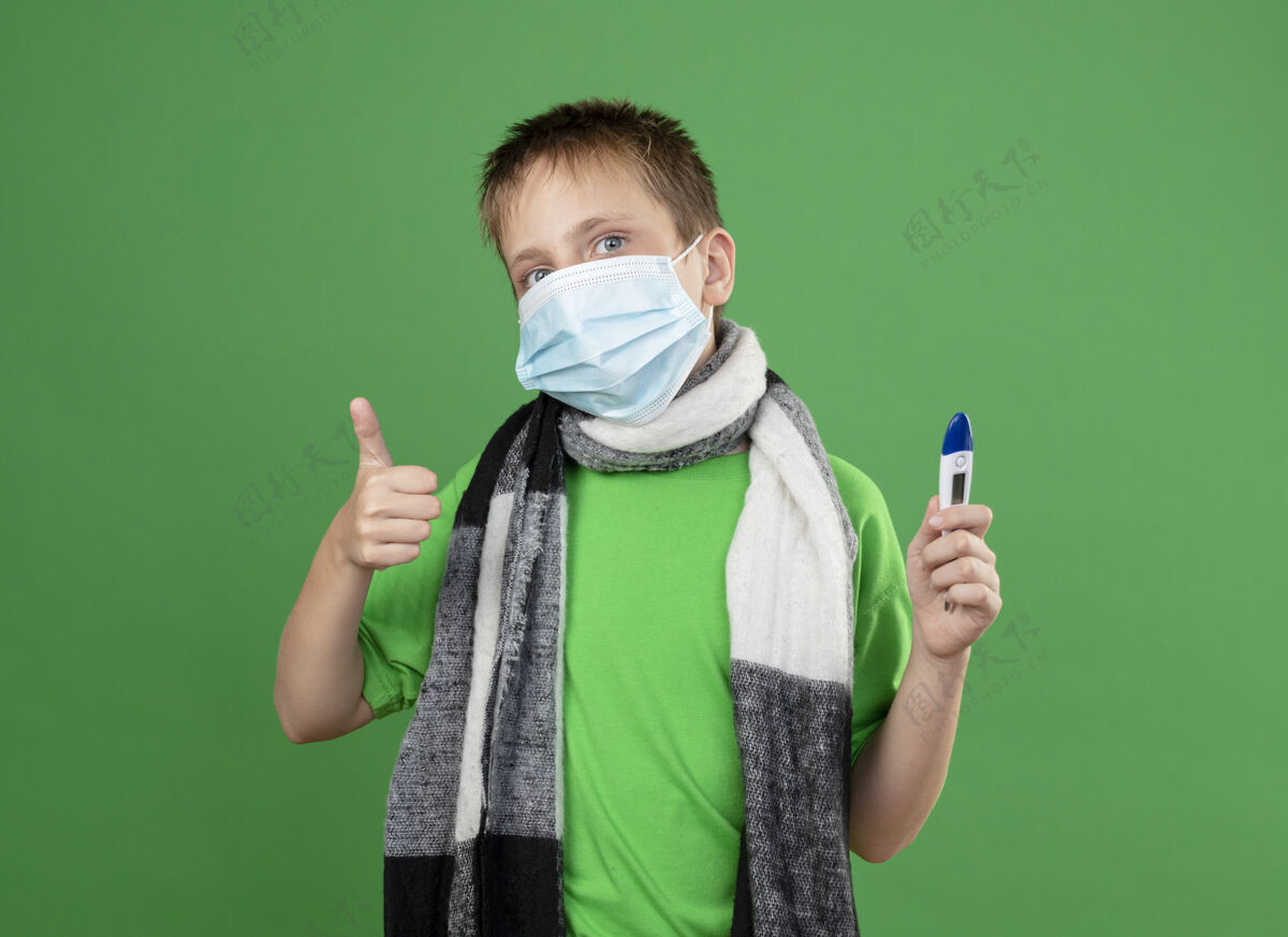 疾病生病的小男孩穿着绿色t恤 脖子上围着暖和的围巾 戴着护面面具 手里拿着温度计 看起来好多了 站在绿色的墙上竖起大拇指面部保护看