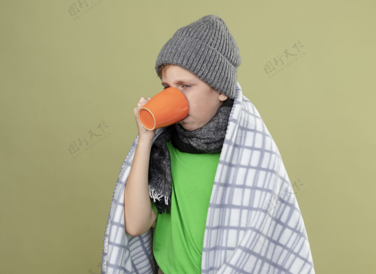 穿生病的小男孩穿着绿色t恤 戴着暖和的围巾和帽子 裹着毯子 喝着热茶 站在轻质的墙上忍受着寒冷帽子热包装