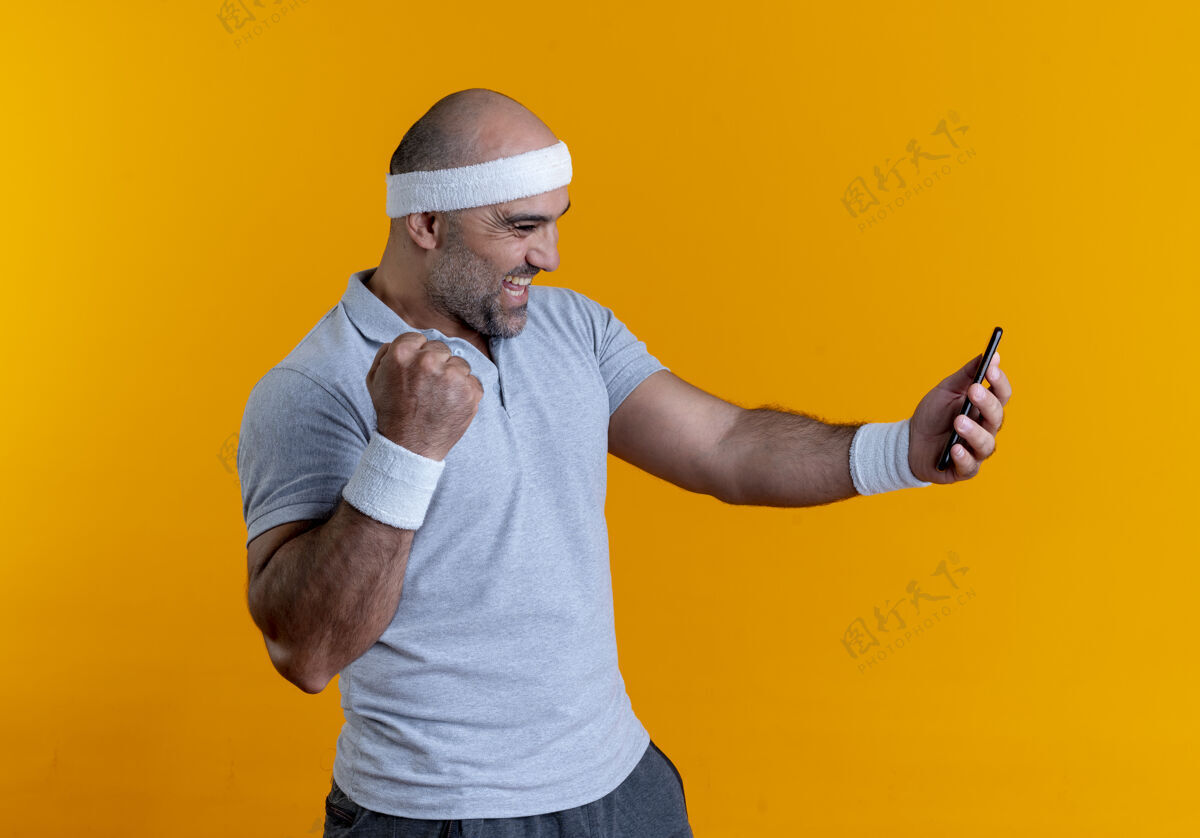 移动戴着头巾的成熟的运动型男人看着手机屏幕 高兴而兴奋地握紧拳头站在橙色的墙上快乐运动男性