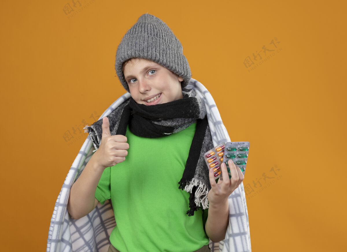 拇指生病的小男孩戴着暖和的帽子 围着围巾裹在毯子里 展示着药片 看起来很好看 微笑着竖起大拇指站在橙色的墙上微笑穿更好
