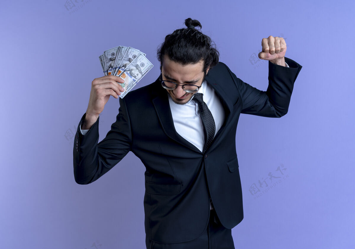 领带一个穿着黑西装戴着眼镜拿着现金的商人站在蓝色的墙上高兴而兴奋地握紧拳头男人眼镜生意