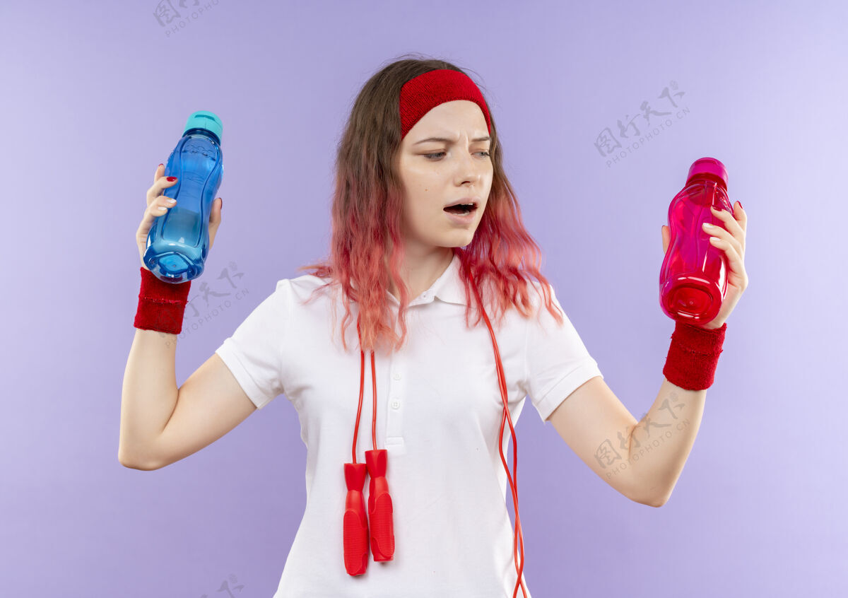 头带戴着头巾 脖子上系着跳绳 手里拿着两瓶水的年轻运动女郎站在紫色的墙上 看上去很困惑 试图做出选择人运动运动装