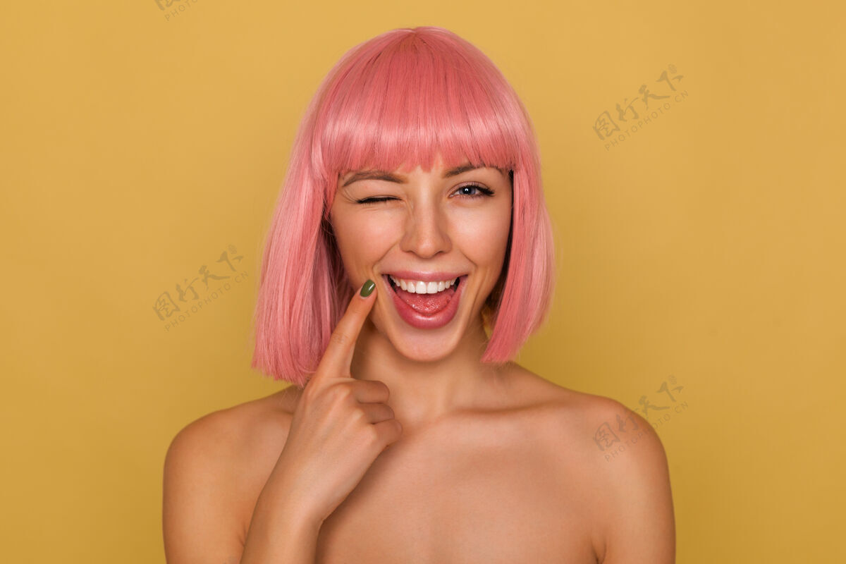 情感快乐的年轻迷人的粉红头发女性 鲍勃的发型把食指放在脸上 面带微笑 愉快地向镜头眨眼 在芥末墙上摆姿势欧洲喜悦特写