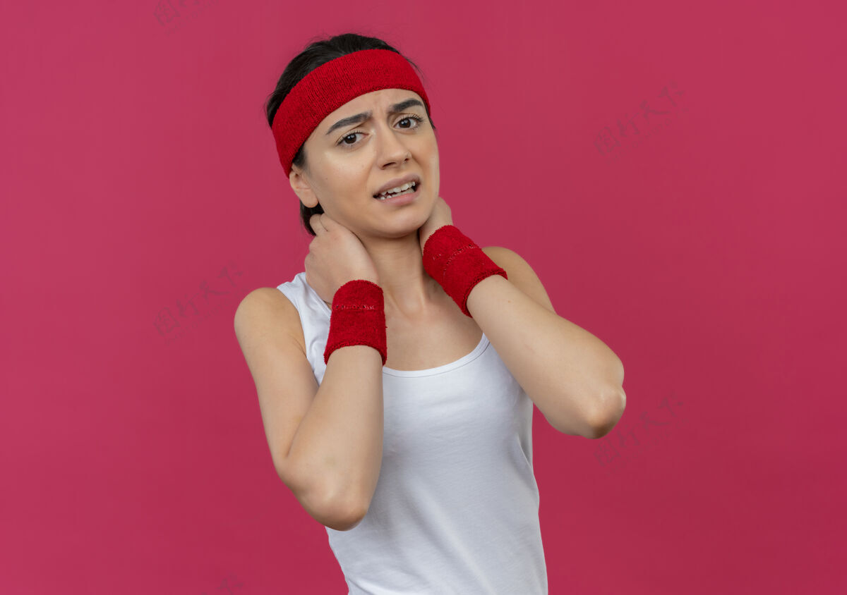 脖子身穿运动服的年轻健身女 头箍贴着脖子 站在粉红色的墙上不高兴地感到疼痛年轻感觉站