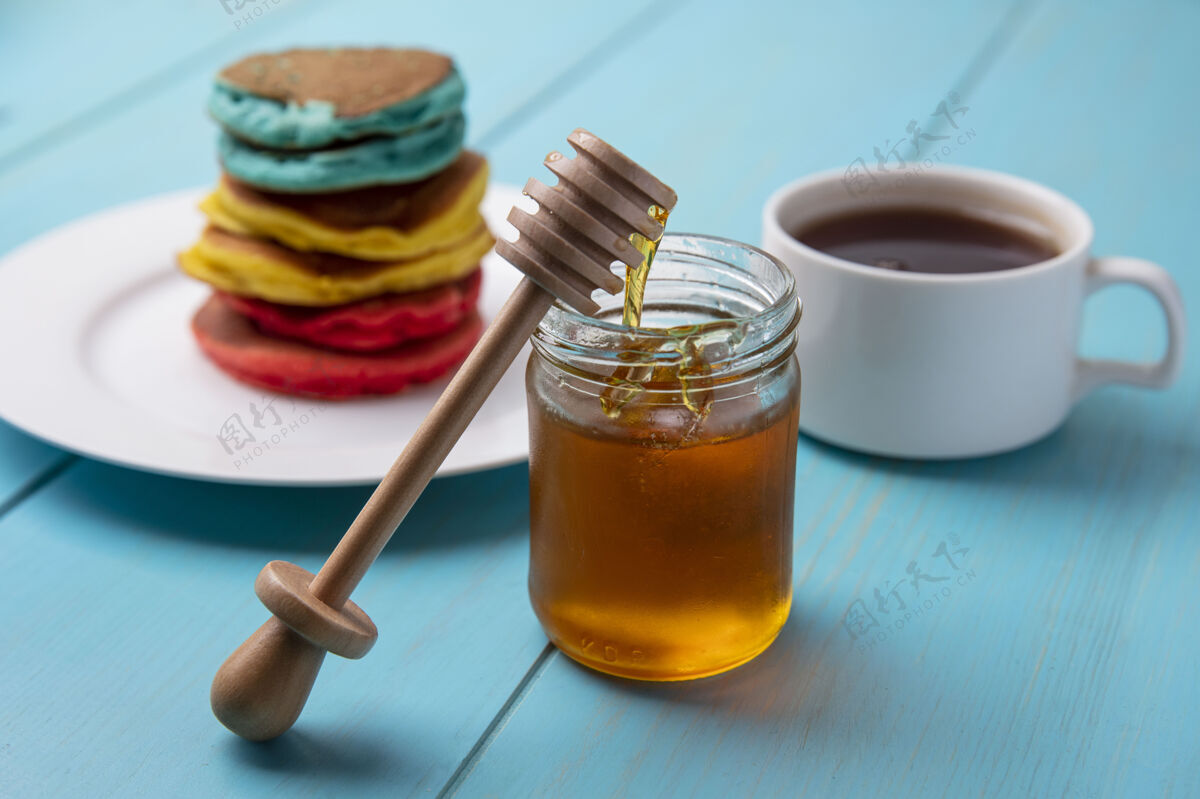 煎饼侧视图五彩缤纷的薄煎饼放在一个盘子里 蜂蜜放在罐子里 木制蜂蜜勺和一杯茶放在绿松石色的背景上勺子风景壁板