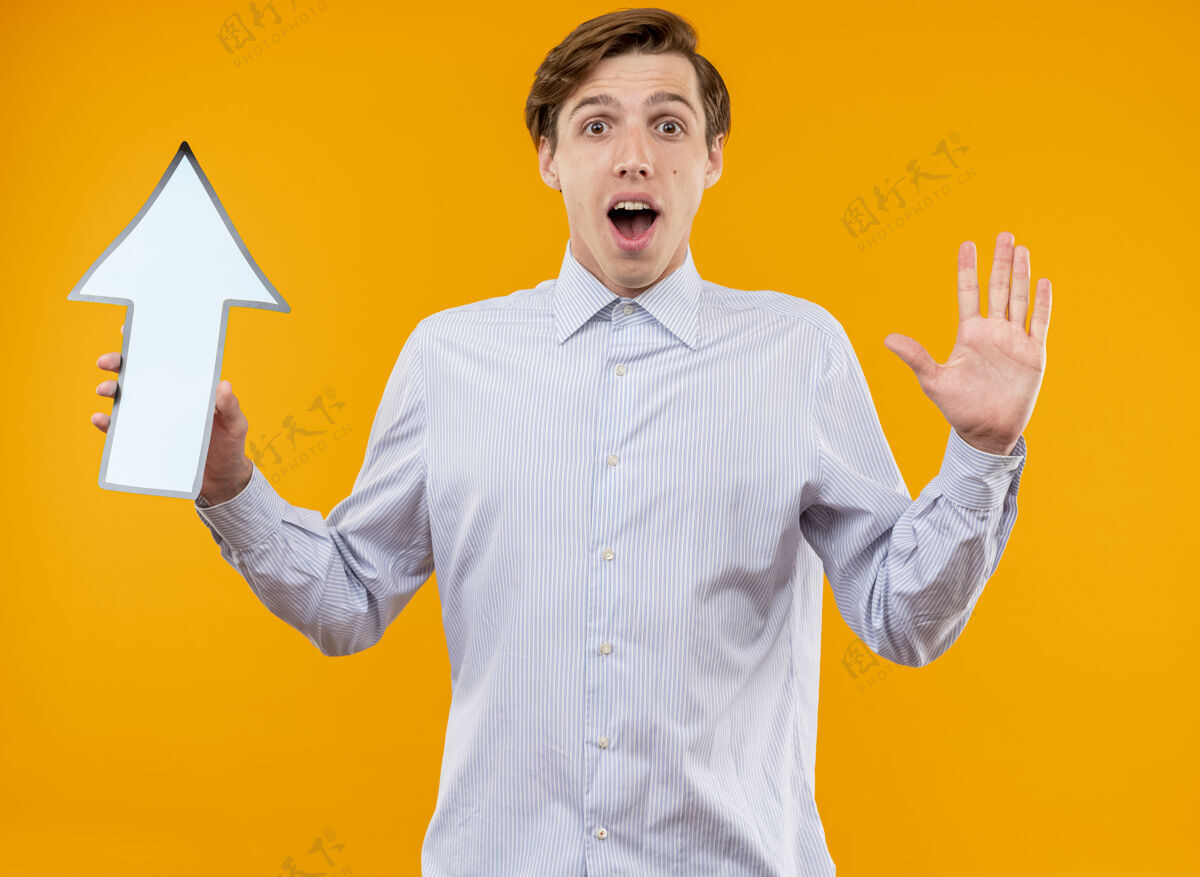 年轻身穿白衬衫的年轻人举着蓝色箭头看着镜头 惊讶而高兴地举起手站在橙色背景上站着手举起