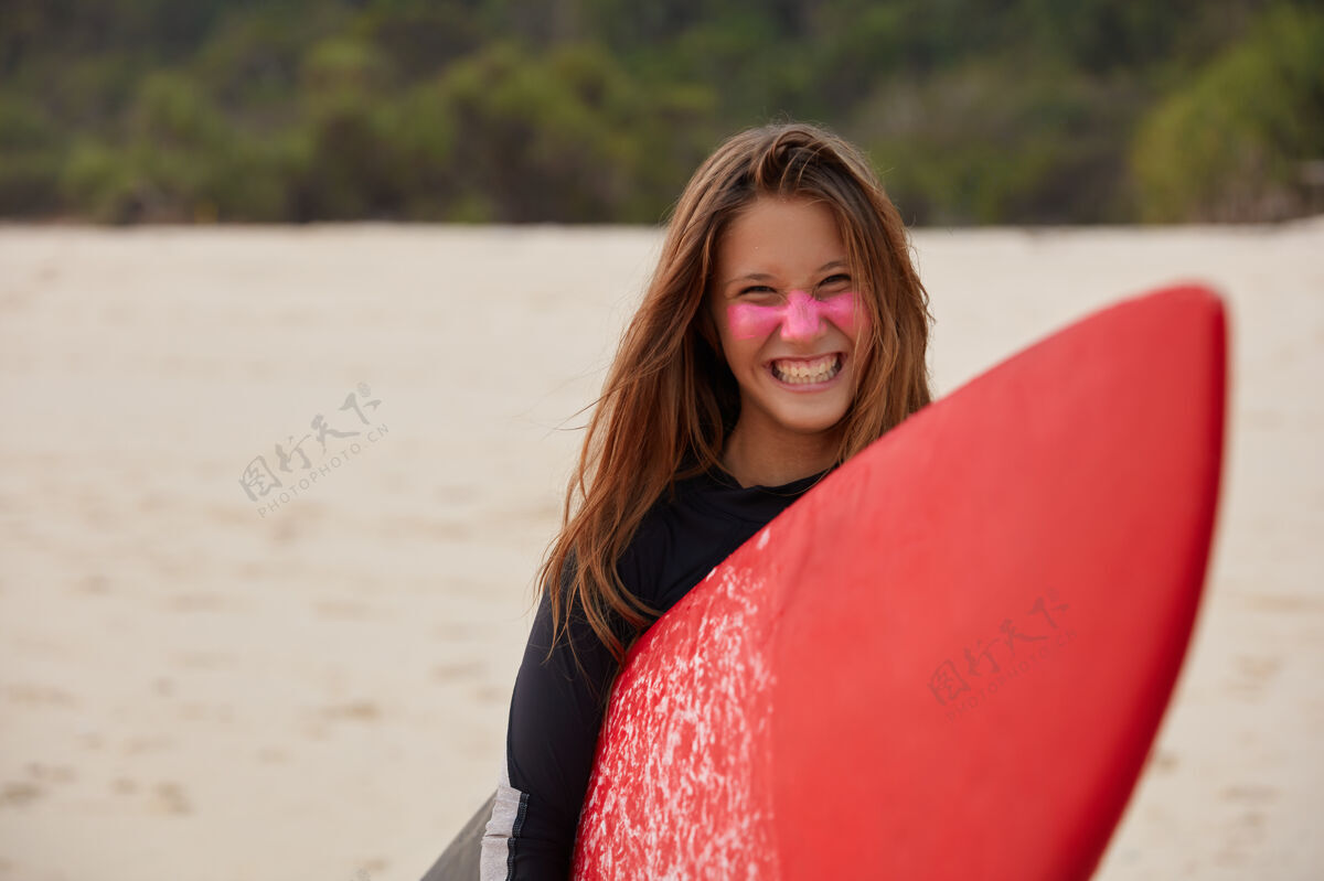 站立开朗的职业年轻女冲浪者笑容灿烂 扛着红蜡冲浪板 脸上有锌 打了大海浪后高兴 靠着沙墙造型 表现积极游泳海滩姿势
