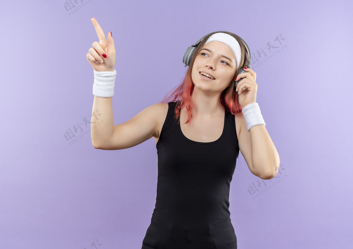耳机身穿运动服的年轻健身女孩头戴耳机 微笑着用手指着站在紫色墙上的一边市民年轻人运动装