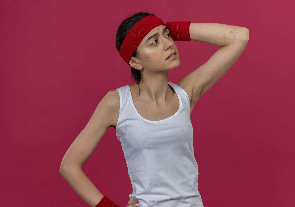 女人身穿运动服的年轻健身女士戴着头巾 站在粉红色的墙上 一边看一边困惑 一边焦急地思考着有疑问的事情健身运动员人