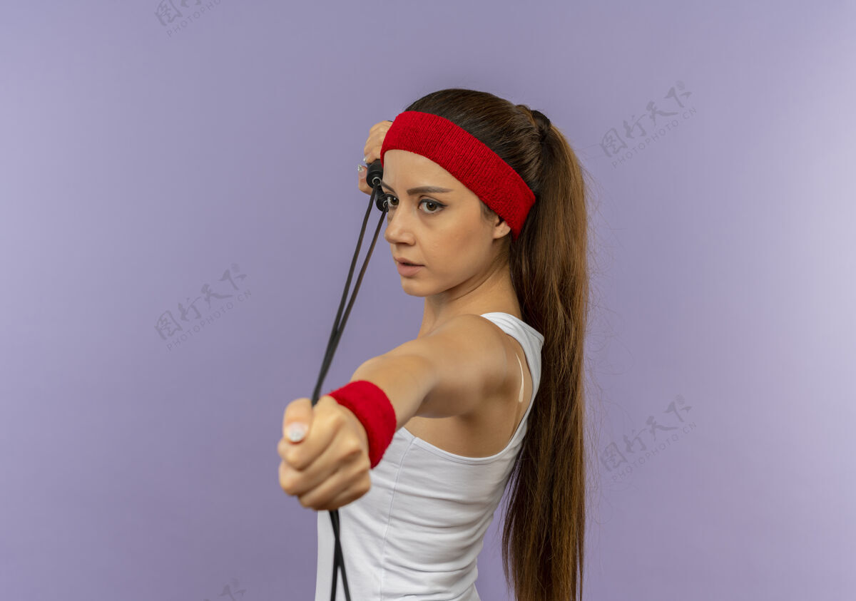 人身穿运动服的年轻健身女 头上戴着头带 拿着跳绳 像是用弓箭瞄准灰色的墙壁运动市民站