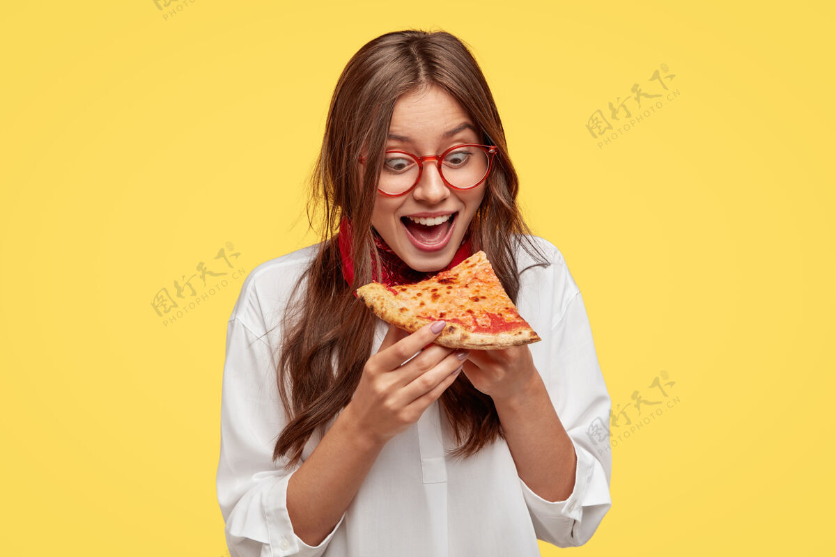 切片饥肠辘辘的学生一边张大嘴巴一边看美味的披萨片 一边想吃 穿着白衬衫 模特靠着黄色的墙壁积极的女人拿着垃圾食品人和吃惊呆了白种人年轻人