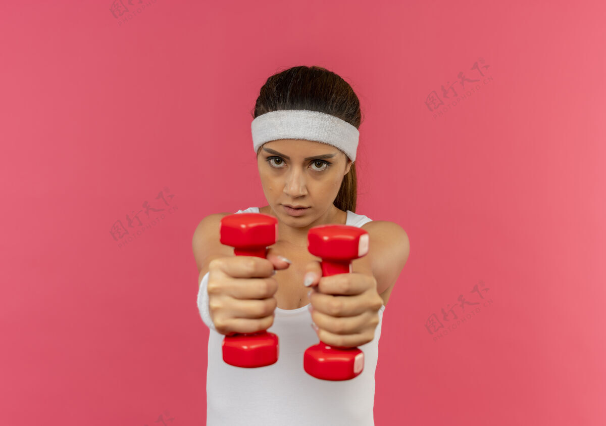 站着身穿运动服 头箍 举着两个哑铃 站在粉色墙壁上做运动的年轻健身女士 看上去很自信健康运动装健身