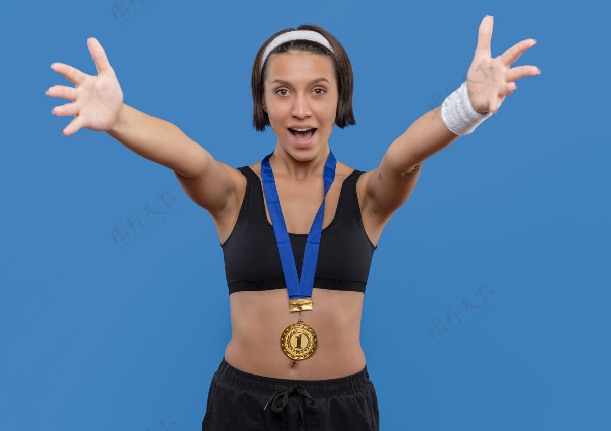 欢迎身穿运动服 脖子上挂着金牌的年轻健身女士站在蓝色的墙上做着欢迎的手势张开双手姿势颈部奖牌